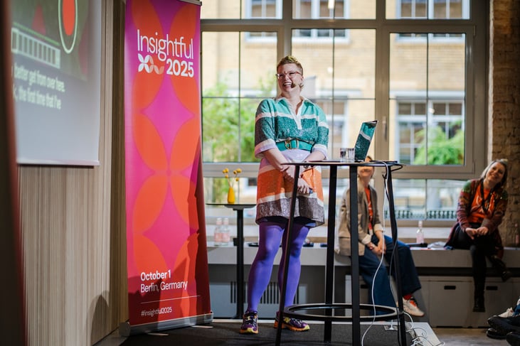 Unn Swanström from Mojang on the stage of Insightful 2025, looking at the screen behind her showing her presentation. She's wearing a green and orange dress, with purple tights and purple sneakers. 